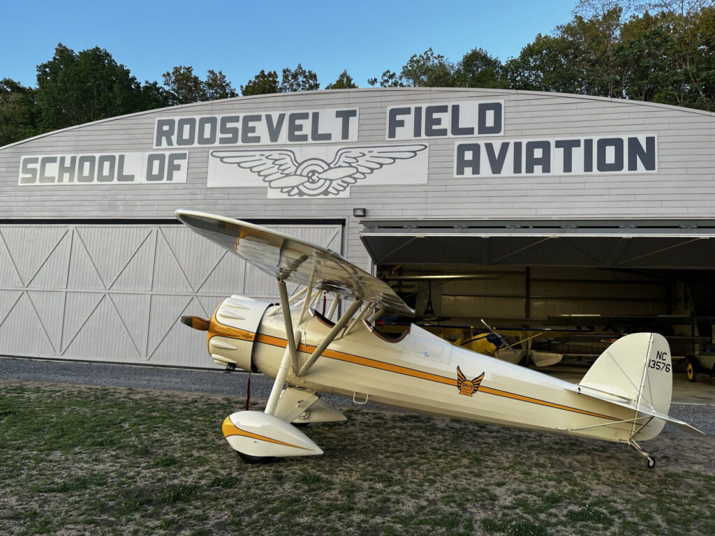 A wide hangar with a curved roof displays the Old Rhinebeck Aerodrome sign, with two yellow planes nearby on grass.