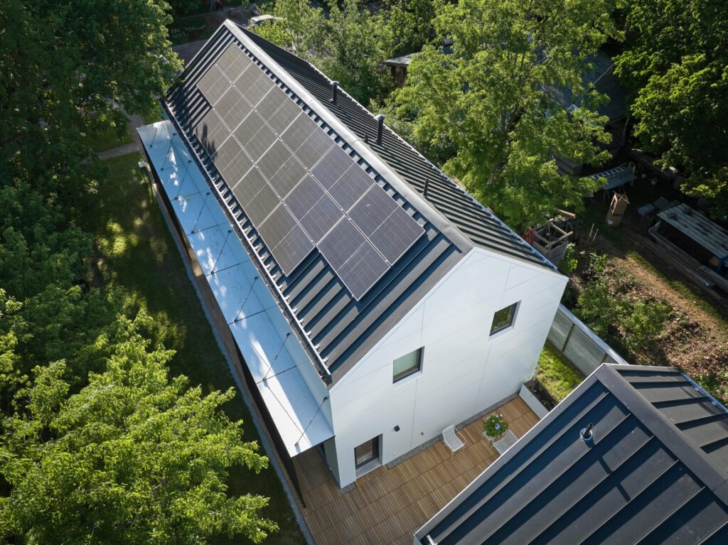 A modern white house with a pitched roof features solar panels and a wood deck, surrounded by trees and greenery.