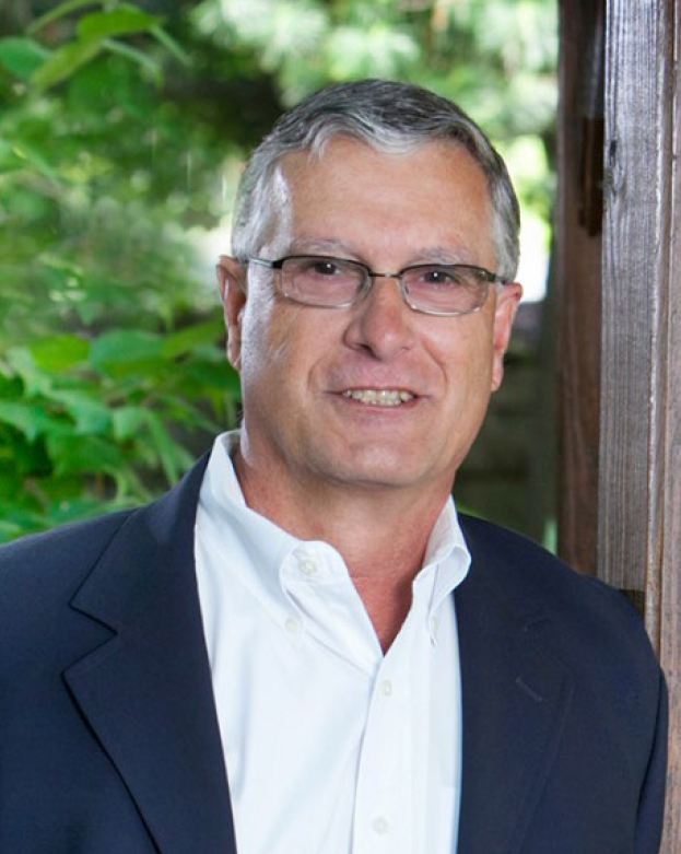A man in a dark blazer and white shirt stands outdoors near a wooden post, smiling with greenery in the background.
