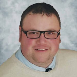 A man wearing thick-rim glasses and wearing a knit vest over a collared shirt smiles at the camera.