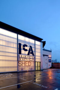 Evening view of the ICA Watershed with its translucent polycarbonate facade glowing from interior light, showcasing the bold logo on the bifold door.