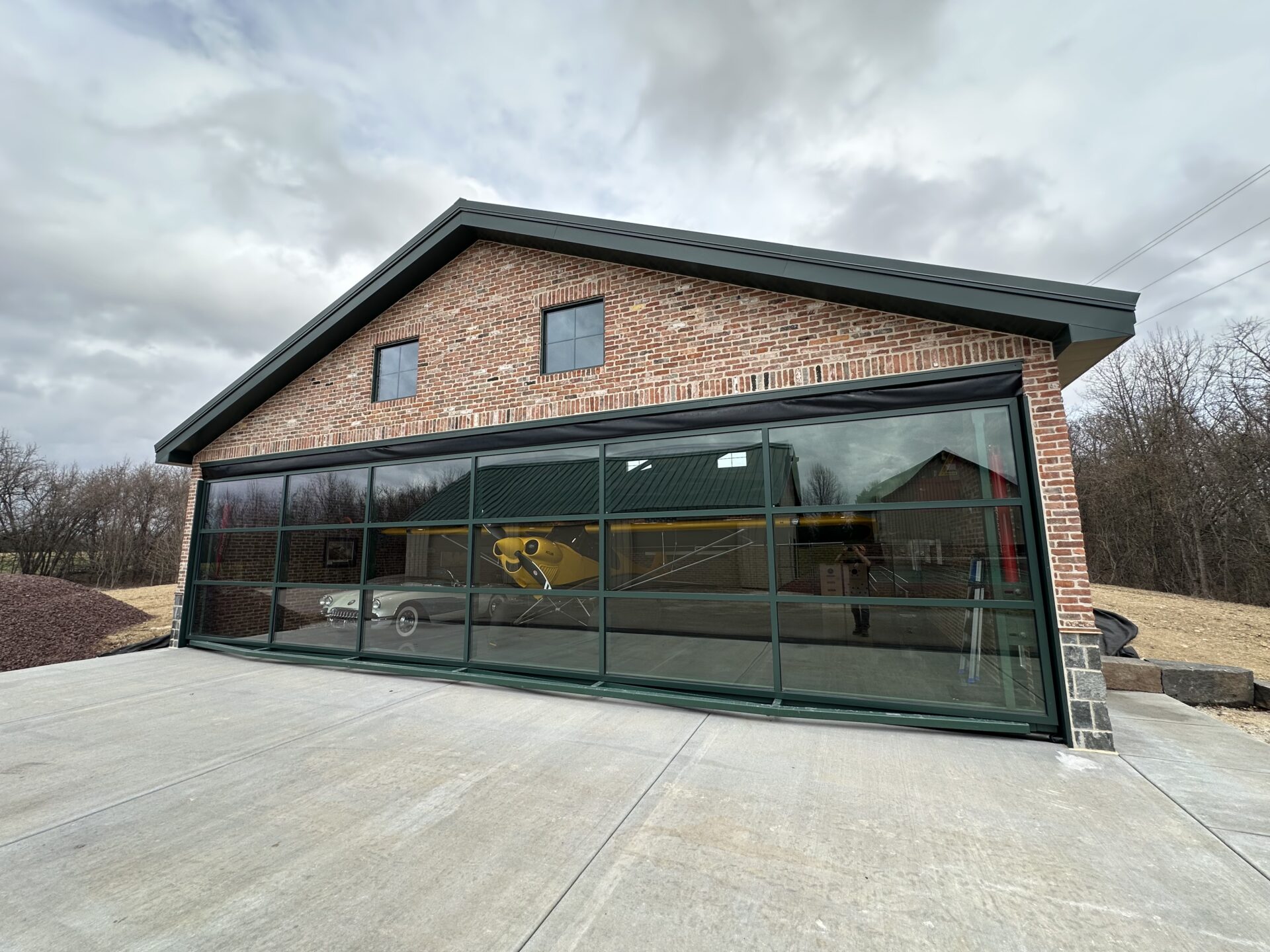 Brick private hangar with a large glass hydraulic door framed in metal, revealing a yellow airplane and car inside under cloudy skies.