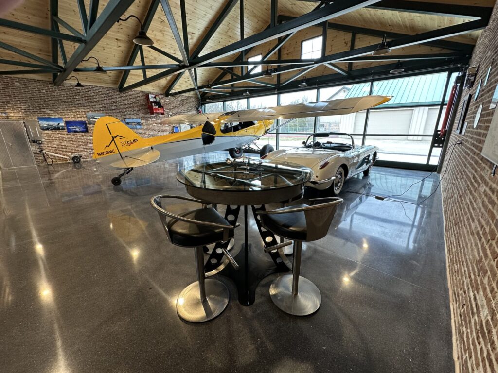 Interior of a private hangar with exposed steel trusses, polished concrete floors, a yellow airplane, classic car, and metal-framed glass table.