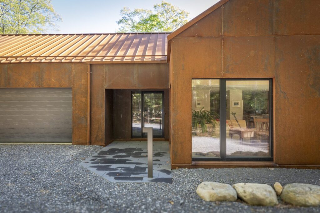 Exterior view of a modern home with rusted steel siding, standing seam metal roof, large glass windows, and stone walkway entrance.