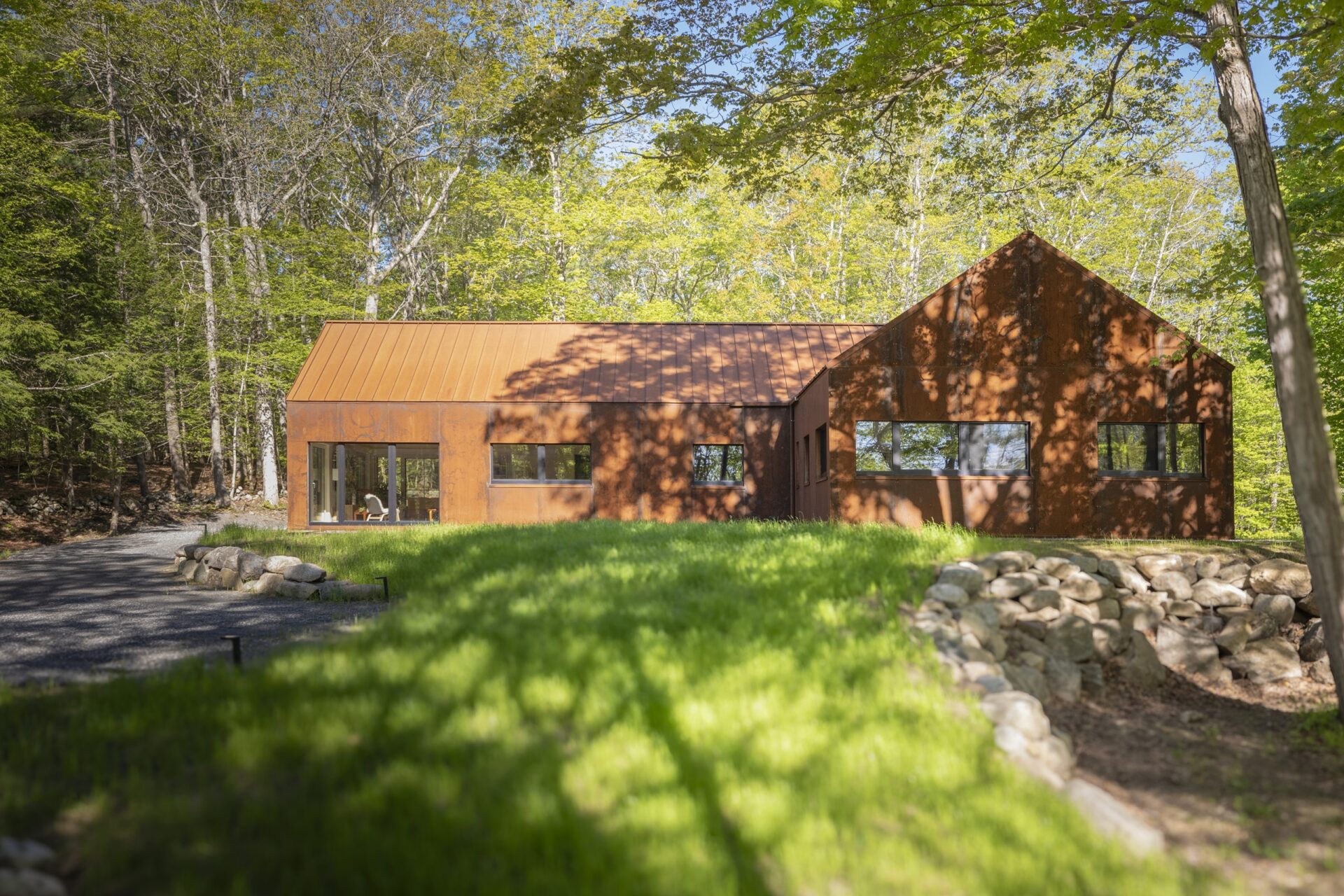 Modern residence clad in rusted steel panels with large windows, set among trees and greenery with a stone-bordered driveway.