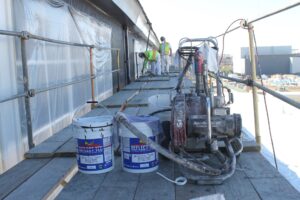 Workers on scaffolding apply protective PVDF coatings to a metal facade, with paint buckets and spray equipment in the foreground.