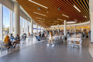 Interior of school with brown metal ceiling. White columns, chairs and tables populate the area. Students sit and walk.