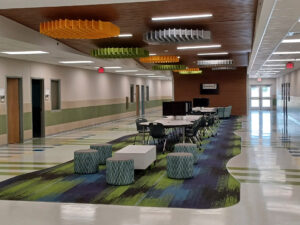School interior hallway with distinct center of ceiling featuring multi-colored geometric light fixtures. A green and blue patterned carpet and small seating stools with short tables run belowthe length and width of the ceiling.
