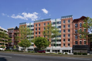 Tall residential building with alternating material pattern on facade. Street and trees in front of the building, and blue sky with some clouds over head