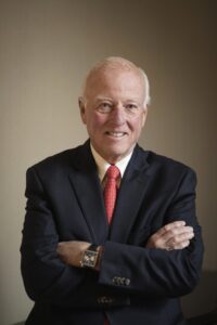 Man in black suit, white dress shirt, and ret necktie crosses arms and smiles in headshot of the donor of a scholarship opportunity for the business of design