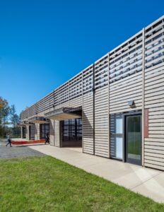 Exterior view of boathouse with outward-opened door. Photographed in the daytime, green grass visible along with people transferring a boat indoors.