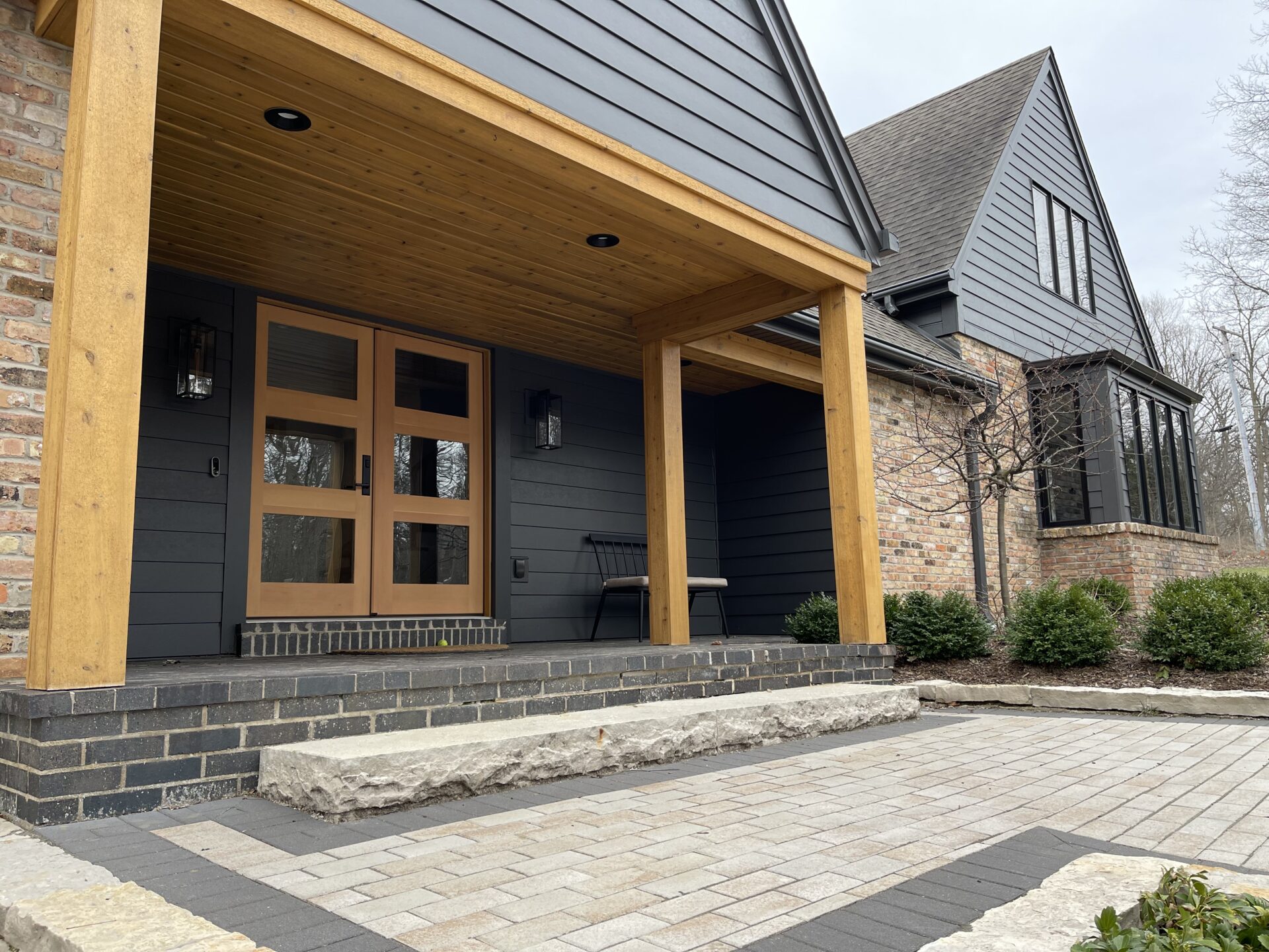 Home facade with camera on a diagonal angle. Black paneling on building exterior is complemented by light brown wooden accents including door frame and pillars.