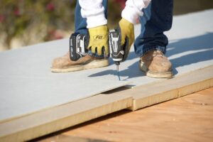 Photograph of polyiso materials being drilled to wood, with worker's gloved hands and work boots visible.