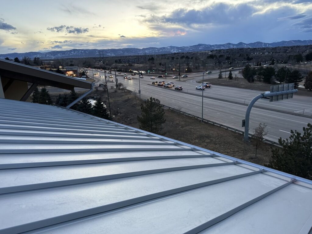 Camera angle: downward from atop a roof. Roof consists of light blue-gray metal panels. Multi-lane highway visible in background.