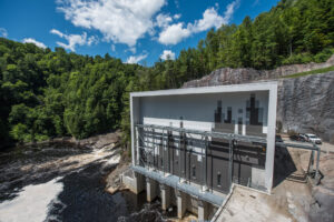 Exterior view of structure mounted on the edge of a rock formation near water surrounded by forestry.