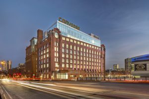 A copper-colored building is positioned on a street corner. Lights glow from the many-windowed facade.