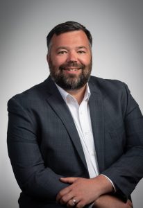 The Architectural Team's New Managing Principal's headshot. He is a man in a suit, smiling in front of a grey background.