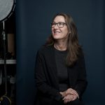 Woman smiles, sitting on stool in front of grey background.