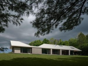 Single-storey residence with a light-colored standing seam metal roof set in a grassy landscape under overcast skies.