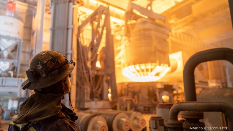 Steelworker in protective gear monitors a glowing molten-metal furnace inside a steel mill.