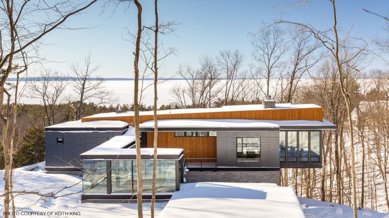 Contemporary multi-level house in a snowy, wooded landscape, featuring dark gray cladding, warm wood accents, large windows, and a glass-fronted entry pavilion.