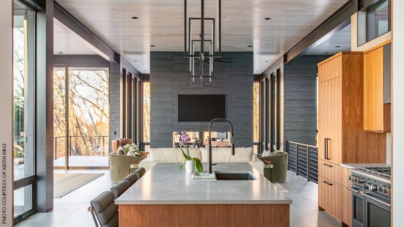 Open-plan modern kitchen and living area with a large wood-and-stone island, tall wood cabinetry, black range, floor-to-ceiling windows, and a dark stone fireplace wall.