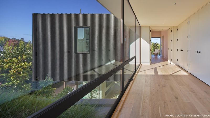 Sunlit modern interior corridor with pale wood flooring, white built-in cupboards, and a full-height glass wall overlooking a dark corrugated exterior façade and trees.