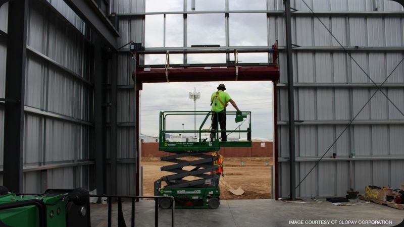 Construction worker on a green scissor lift installing a large overhead sectional door inside a steel-framed industrial building.