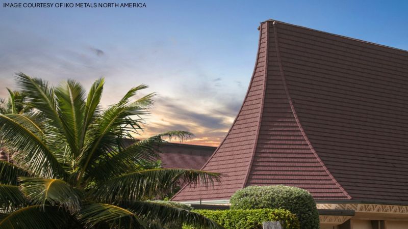 A steep, dark red roof with a tall, curved tower-like section, framed by palm trees and tropical greenery at sunset.