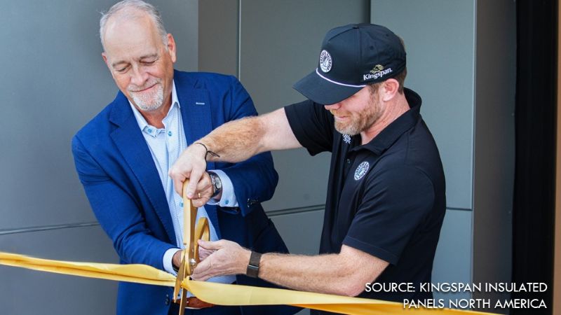 Two men work together to cut a wide yellow ribbon with oversized ceremonial scissors. One wears a blue suit jacket, while the other wears a black polo shirt and baseball cap.