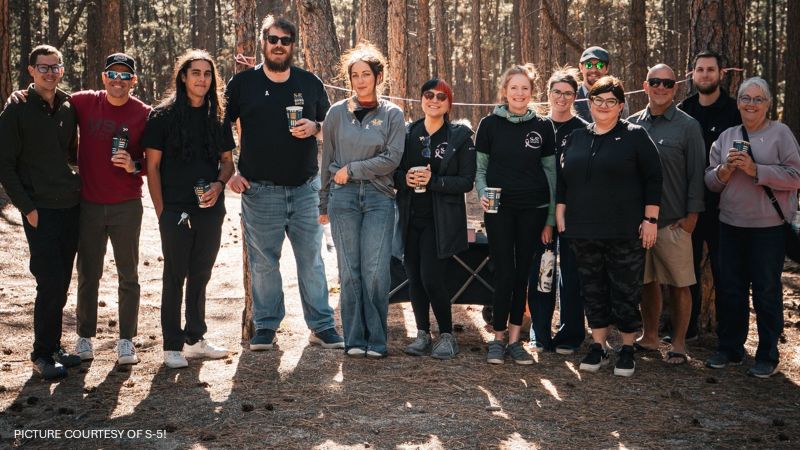 A group of S-5! employees stand together outdoors among trees, several holding cups.