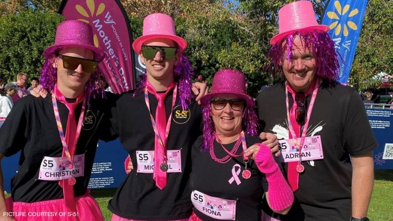 Four participants wear pink hats and wigs, ribbons and event medals at a breast cancer fundraiser.