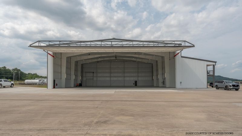 Exterior: Wide-open aircraft hangar with a large bifold door, concrete apron, and cloudy sky overhead.